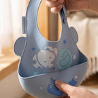 Marcus & Marcus pastel blue silicone baby bib with a handle, decorated with cute cartoon animal and star patterns, being held by hands in a softly lit indoor setting.