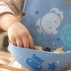 A close-up of a child's hand reaching into Marcus & Marcus pastelblue silicone bib wide catch decorated with cartoon animal and star illustrations. 