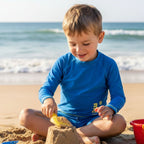 Child wearing long sleeve rash guard with marckids logo building a sandcastle on a beach with ocean in the background