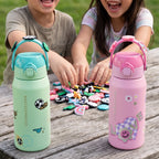 Two kids sitting at a wooden picnic table outdoors, smiling and playing with small colorful items. In the foreground, two insulated water bottles are shown: one mint green with a turquoise lid and handle, and one pink with a pink lid and handle.