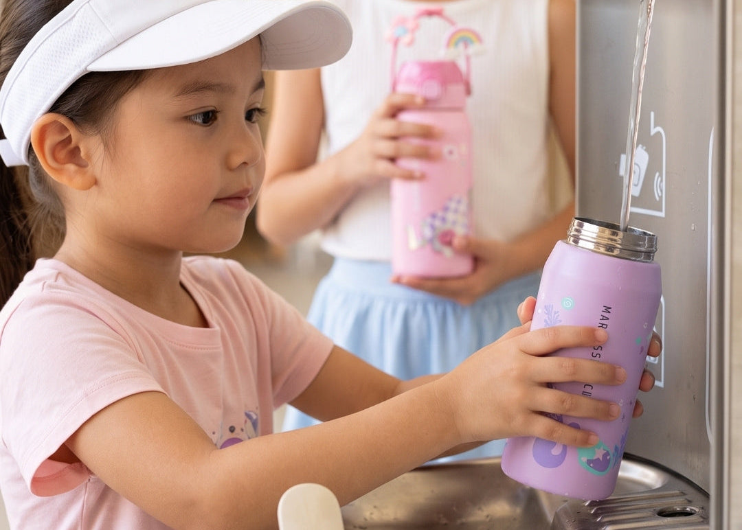 A young girl in a pink shirt and white visor fills a lavender insulated water bottle from a stainless steel water dispenser, with another child holding a pink bottle in the background.
