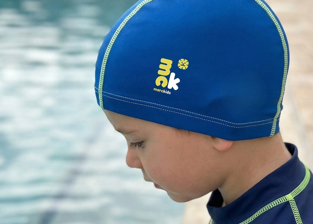 Child wearing marckids blue swim cap with a brand logo, standing by a pool.