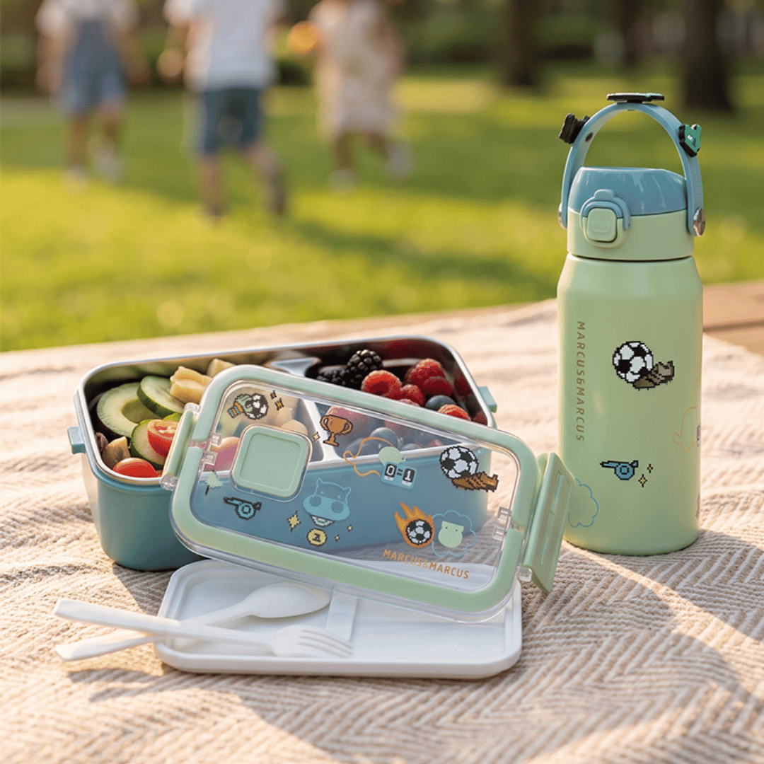 A light green and white kids' lunch set with a reusable bottle and bento box, styled on a picnic blanket in a sunny park with children blurred in the background.